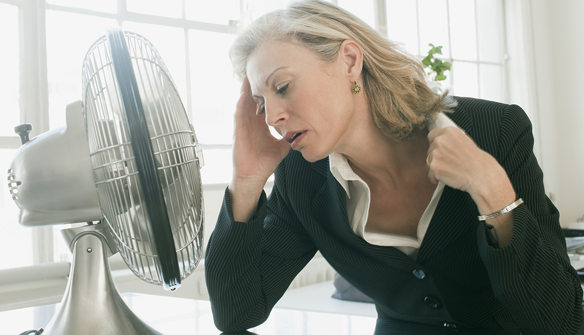 Mature woman in a business suit sitting in front of electric fan, looking uncomfortably hot.